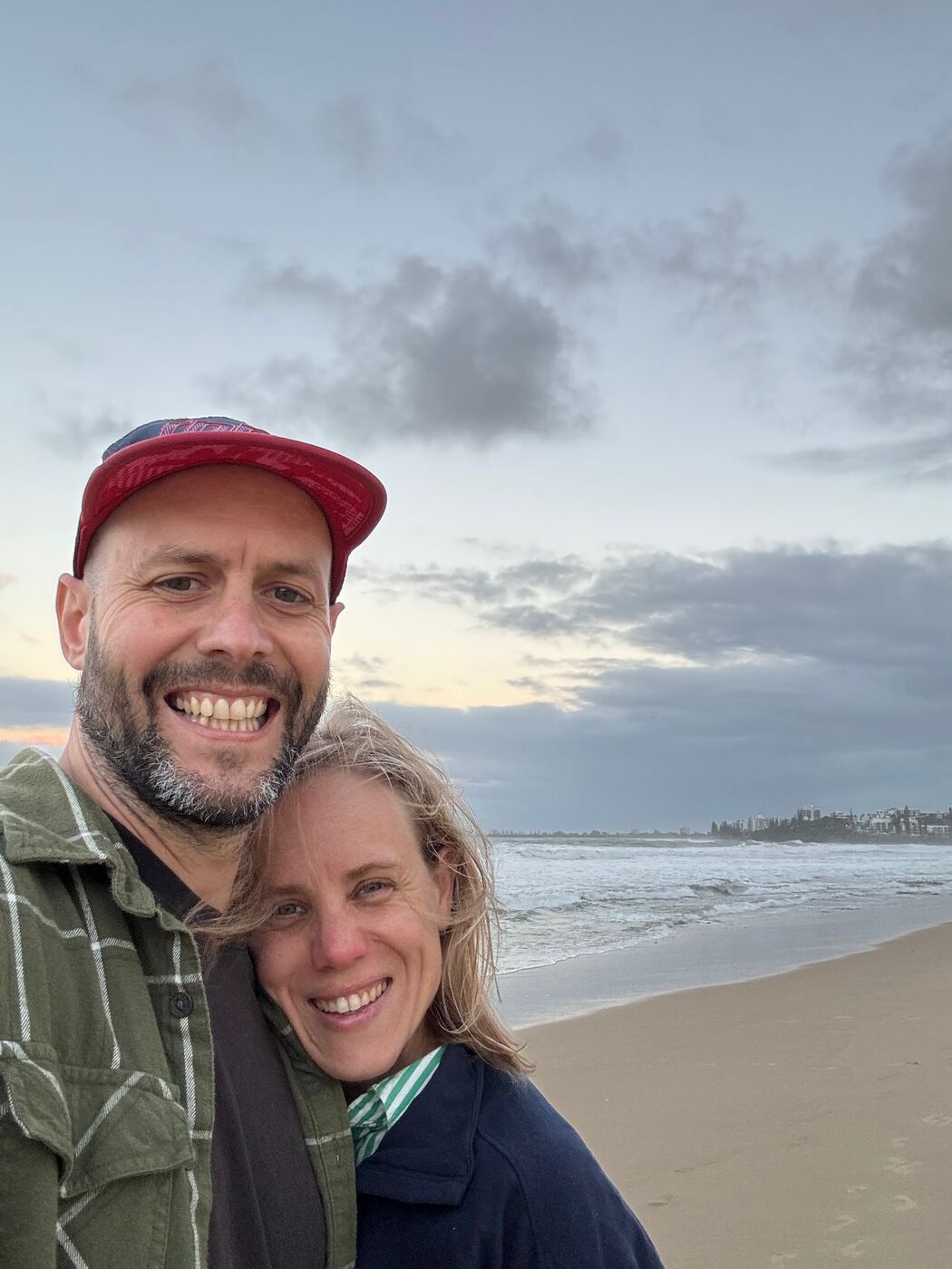 Jess and Greg cheek to cheek on a beach at dusk, with the surf behind them.