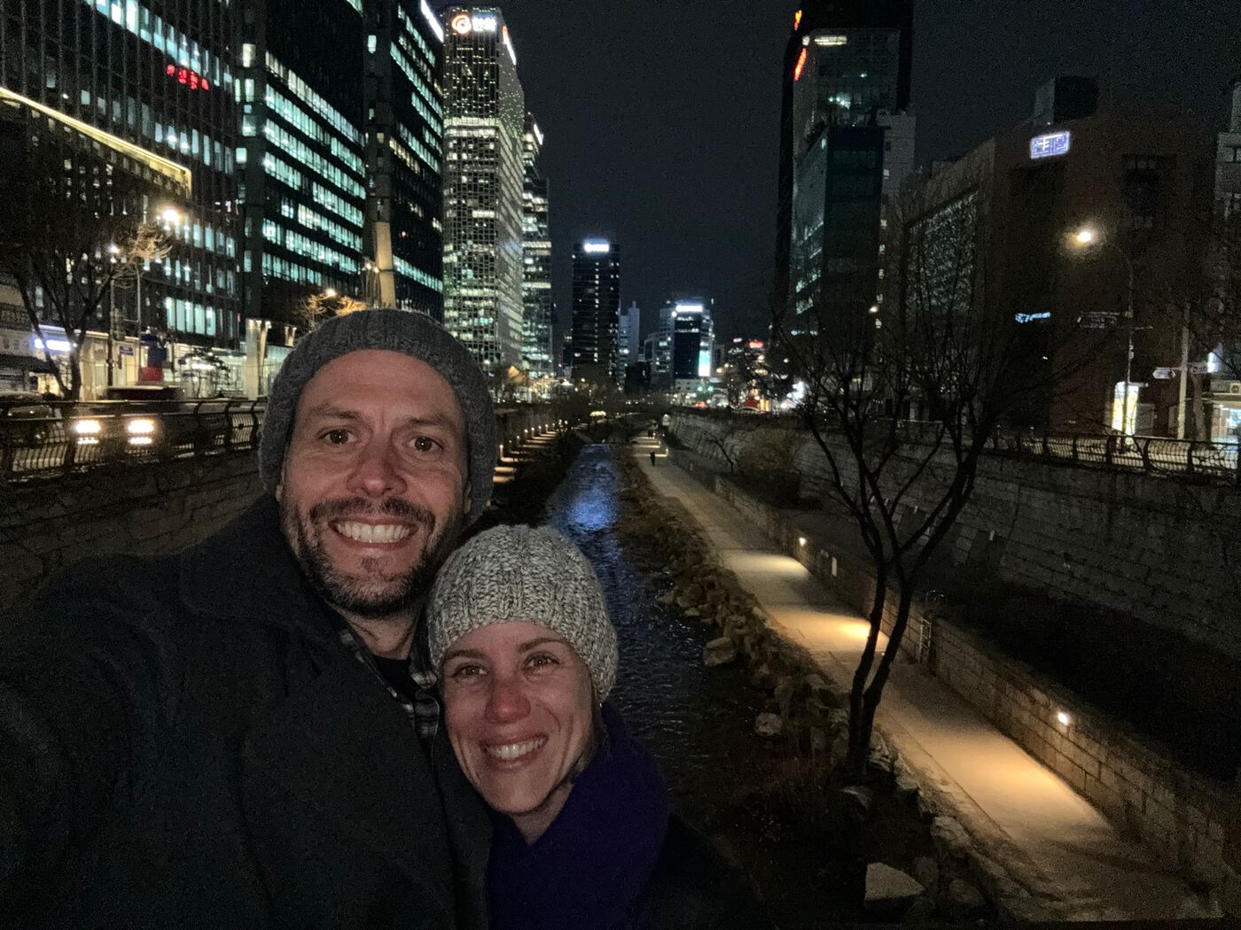 Jess and Greg in woollen hats beside a lit canal at night, with city towers behind them.