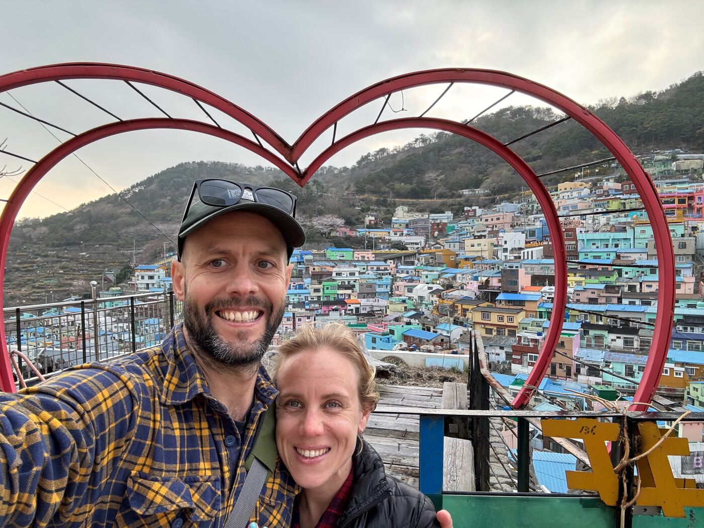 Jess and Greg inside a large red heart frame, with a hillside of coloured houses behind them.