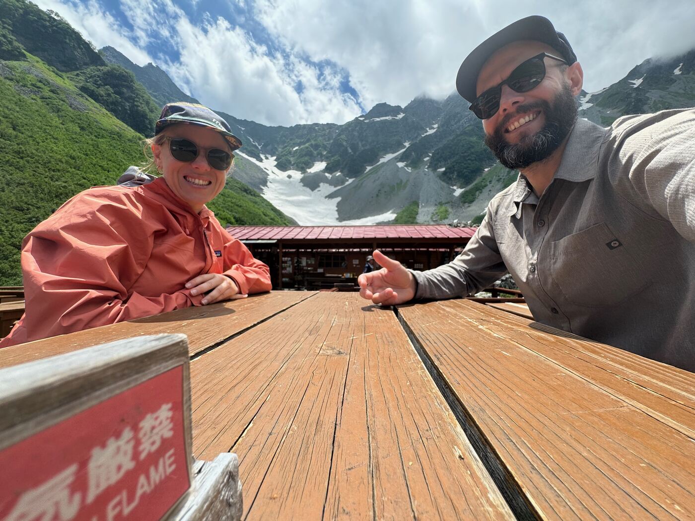 Jess and Greg at a wooden table outside a mountain hut, below a snow-streaked ridge.