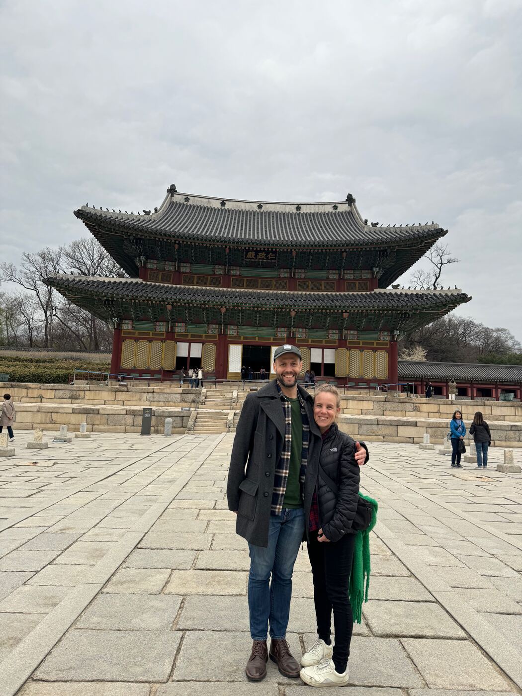 Jess and Greg in winter coats on a stone courtyard, in front of a tiled palace hall.