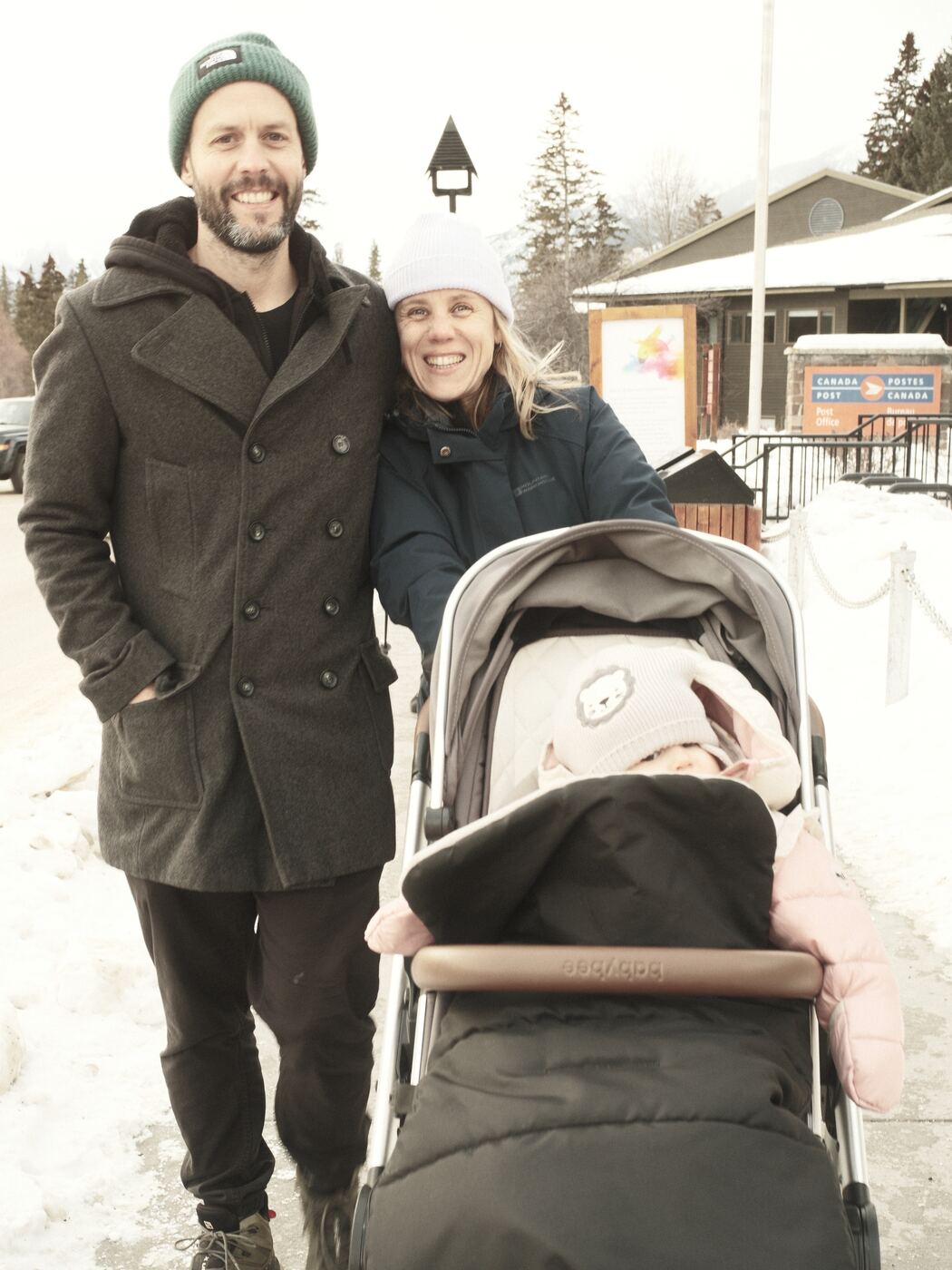 Jess and Greg with a pram on a snowy road, both in winter coats.