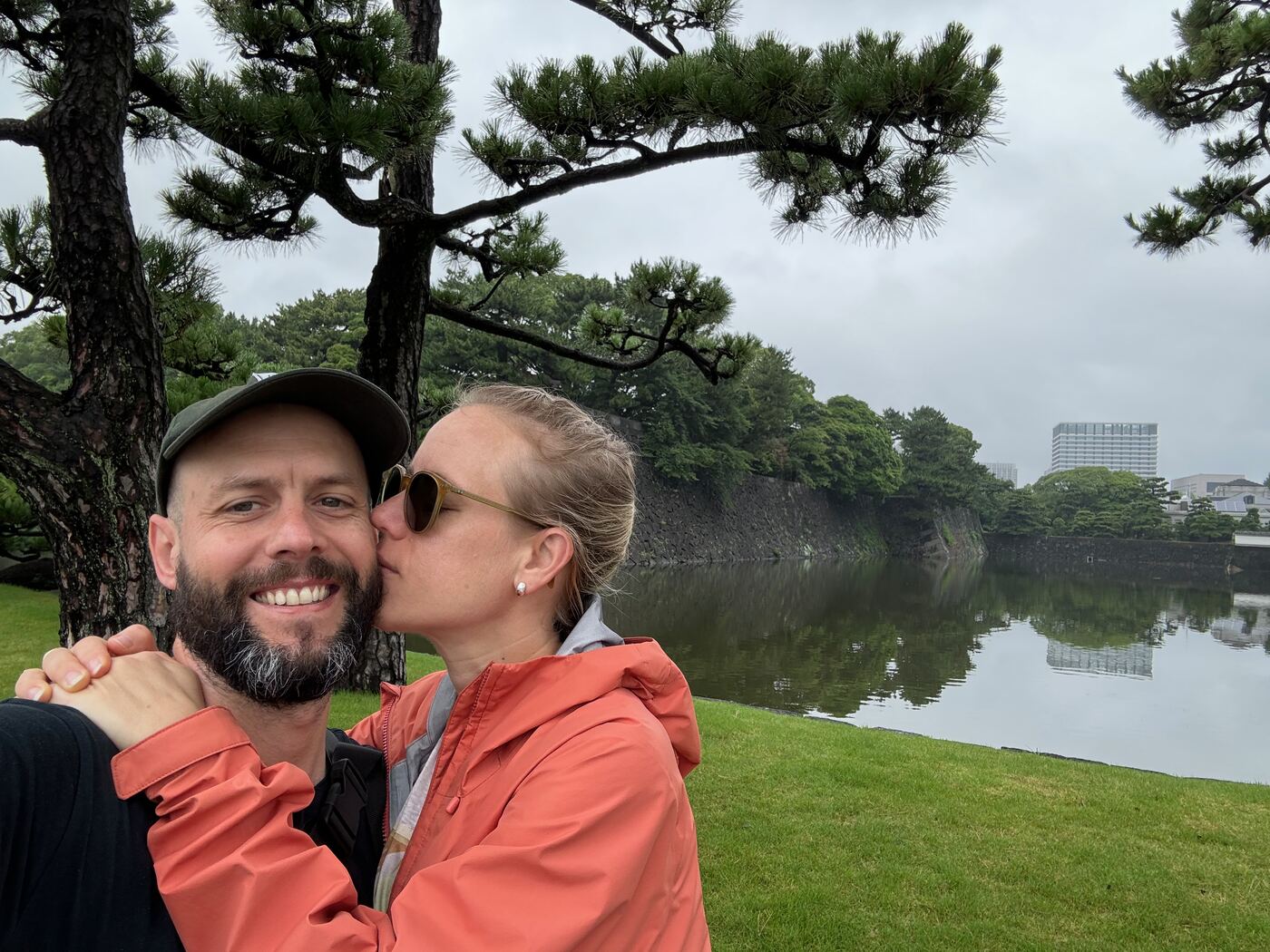Jess gives Greg a kiss on the cheek beside a still moat, under a pine tree.