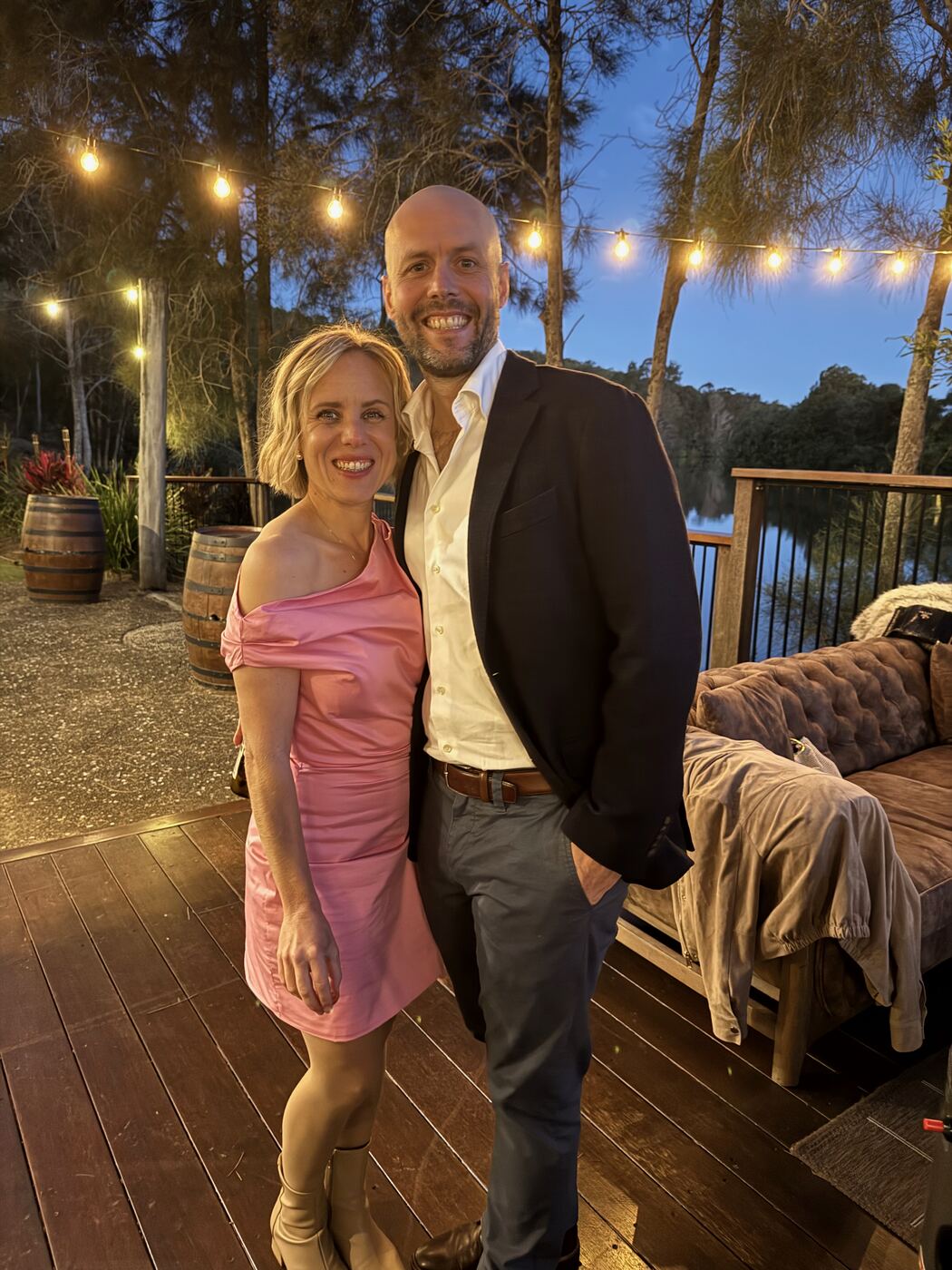 Jess and Greg dressed for a party at dusk, with string lights on the deck behind them.